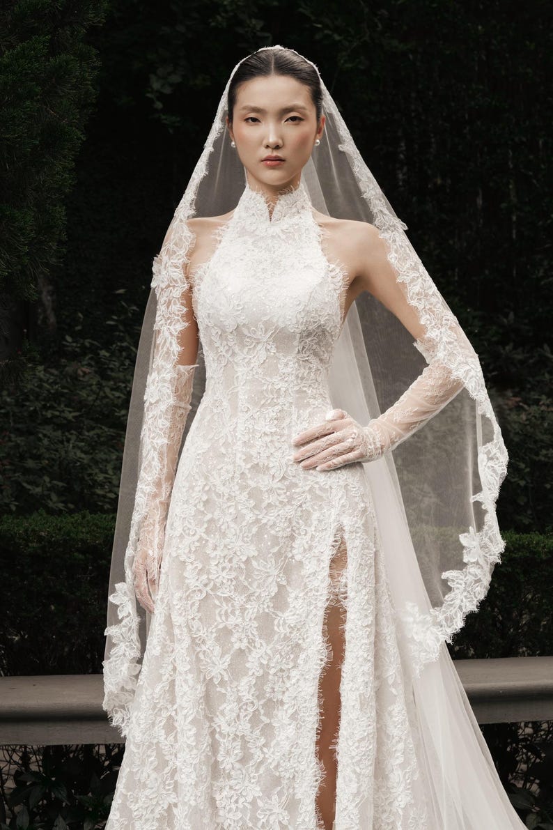 May include: A white lace wedding dress with a high neckline and a thigh-high slit. The dress features long, sheer sleeves and a matching veil with lace trim. The bride is wearing white gloves and pearl earrings.