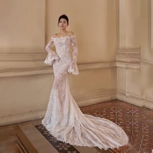 Puede incluir: Un vestido de novia blanco, con hombros descubiertos, con mangas largas y una silueta de sirena. El vestido presenta intrincados detalles de encaje y una larga cola. Las mangas tienen un diseño acampanado. El vestido es largo hasta el suelo y parece estar hecho de una tela delicada.