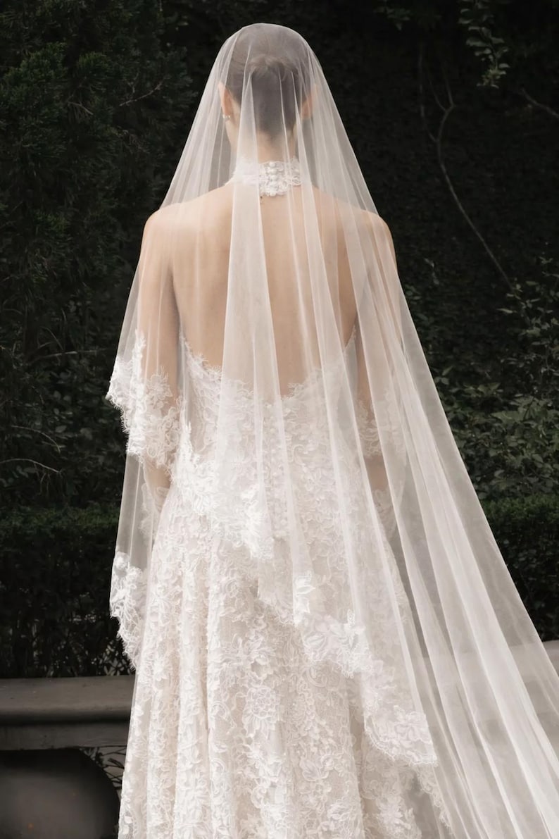 May include: A white lace wedding dress with a long, flowing veil. The dress features intricate lace detailing and a high neckline. The veil cascades down the back, creating an elegant and romantic look.
