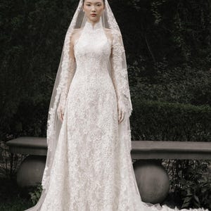 May include: A white lace wedding dress with a high neckline and long sleeves. The dress features intricate floral lace detailing and a flowing skirt. A matching lace veil drapes from the head, creating an elegant bridal look.