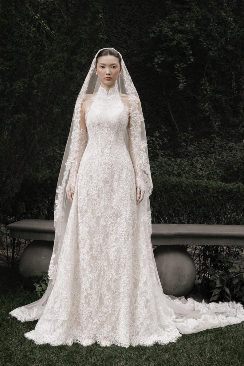 May include: A white lace wedding dress with a high neckline and long sleeves. The dress features intricate floral lace detailing and a flowing skirt. A matching lace veil drapes from the head, creating an elegant bridal look.