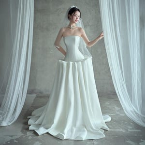 Peut inclure: Une robe de mariée blanche sans bretelles avec un corsage ajusté et une jupe ample. La robe présente un élément de design unique à la taille, créant une silhouette distinctive. Un voile transparent et un délicat coiffe complètent le look de mariée.