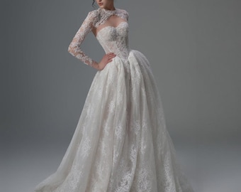 Pode incluir: Um vestido branco longo até ao chão com um corpete cai-cai em forma de coração com sobreposição de renda intrincada, uma única manga comprida de renda e uma saia volumosa com padrões florais de renda detalhados do corpete até à bainha.