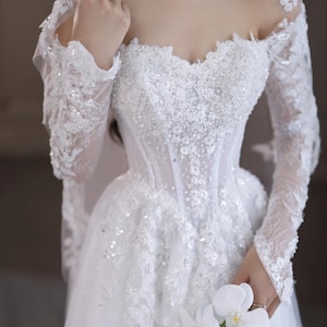 May include: A white wedding dress with a sweetheart neckline, long lace sleeves, and a full skirt. The bodice is embellished with sequins and beads. A bouquet of white flowers is held in the hand.