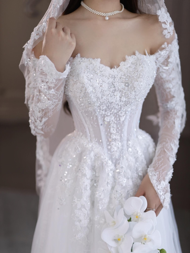 May include: A white wedding dress with a sweetheart neckline, long lace sleeves, and a full skirt. The bodice is embellished with sequins and beads. A bouquet of white flowers is held in the hand.