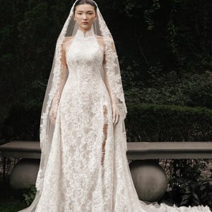 May include: A white lace wedding dress with a high neckline and long sleeves. The dress features a thigh-high slit and a long train. A matching lace veil is draped over the head and shoulders. The dress is worn outdoors.