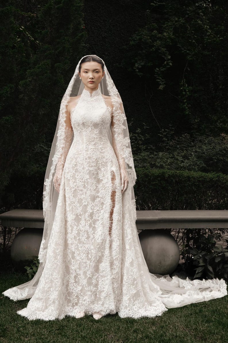 May include: A white lace wedding dress with a high neckline and long sleeves. The dress features a thigh-high slit and a long train. A matching lace veil is draped over the head and shoulders. The dress is worn outdoors.