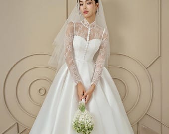Puede incluir: Un vestido de novia de satén blanco con cuello alto y mangas largas de encaje. El vestido presenta un corpiño abotonado y una falda amplia. Un velo transparente y un ramo de flores blancas completan el look nupcial.