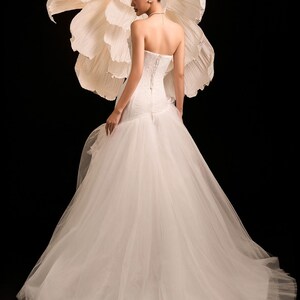May include: A white bridal gown with a strapless bodice and a corset back, paired with a flowing tulle skirt. A large, cream-coloured paper flower is behind the dress. The background is black.
