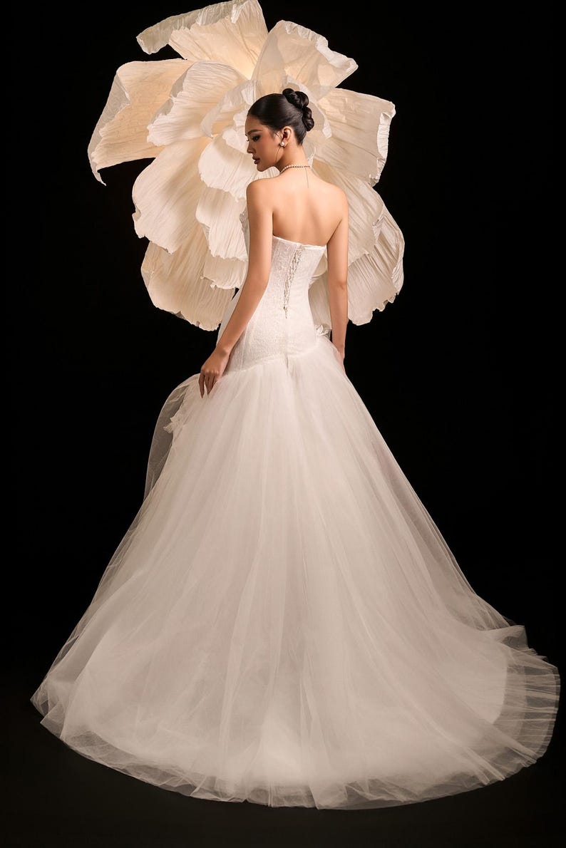 May include: A white bridal gown with a strapless bodice and a corset back, paired with a flowing tulle skirt. A large, cream-coloured paper flower is behind the dress. The background is black.