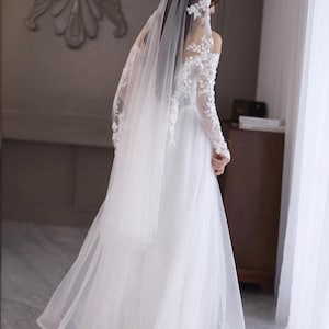 May include: A white wedding dress with a long, flowing skirt and a sheer veil. The dress features delicate lace detailing on the sleeves and bodice. The bride is standing near a window, with the veil cascading down her back.