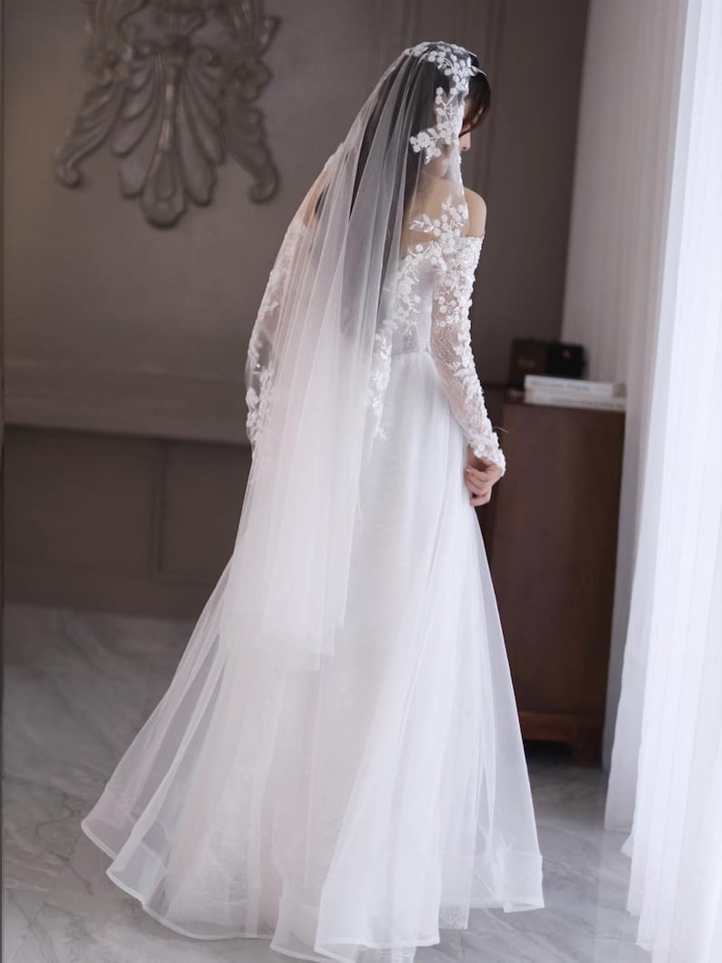 May include: A white wedding dress with a long, flowing skirt and a sheer veil. The dress features delicate lace detailing on the sleeves and bodice. The bride is standing near a window, with the veil cascading down her back.