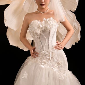 May include: A white bridal gown with floral detailing and a tulle skirt. The strapless bodice has beadwork and a corset design. A pearl necklace and veil complete the look. A large, white paper flower is in the background.