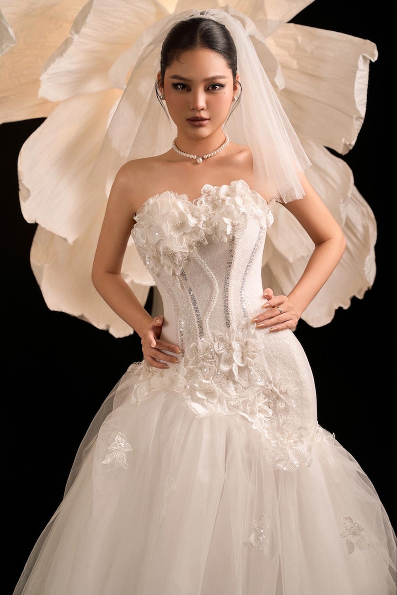 May include: A white bridal gown with floral detailing and a tulle skirt. The strapless bodice has beadwork and a corset design. A pearl necklace and veil complete the look. A large, white paper flower is in the background.