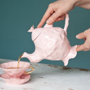 Peut inclure: Une théière rose avec des finitions dorées verse du thé dans une tasse à thé et une soucoupe roses. La théière et la tasse à thé ont une surface texturée et bosselée. Il y a un sucrier rose assorti avec des finitions dorées en arrière-plan.