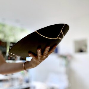 Puede incluir: Un cuenco de cer&aacute;mica negra con l&iacute;neas doradas de reparaci&oacute;n kintsugi. El cuenco se sostiene en &aacute;ngulo, mostrando las grietas reparadas. Las l&iacute;neas doradas a&ntilde;aden un elemento decorativo, resaltando la belleza de la imperfecci&oacute;n. Una pieza de arte &uacute;nica.