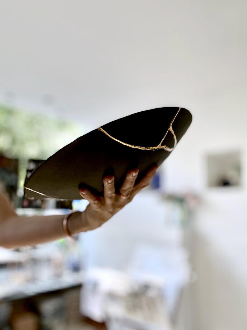 Puede incluir: Un cuenco de cer&aacute;mica negra con l&iacute;neas doradas de reparaci&oacute;n kintsugi. El cuenco se sostiene en &aacute;ngulo, mostrando las grietas reparadas. Las l&iacute;neas doradas a&ntilde;aden un elemento decorativo, resaltando la belleza de la imperfecci&oacute;n. Una pieza de arte &uacute;nica.