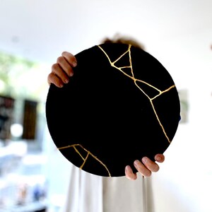 Puede incluir: Una pieza decorativa redonda negra con l&iacute;neas doradas al estilo kintsugi. La pieza est&aacute; sostenida por dos manos, y el fondo est&aacute; borroso.