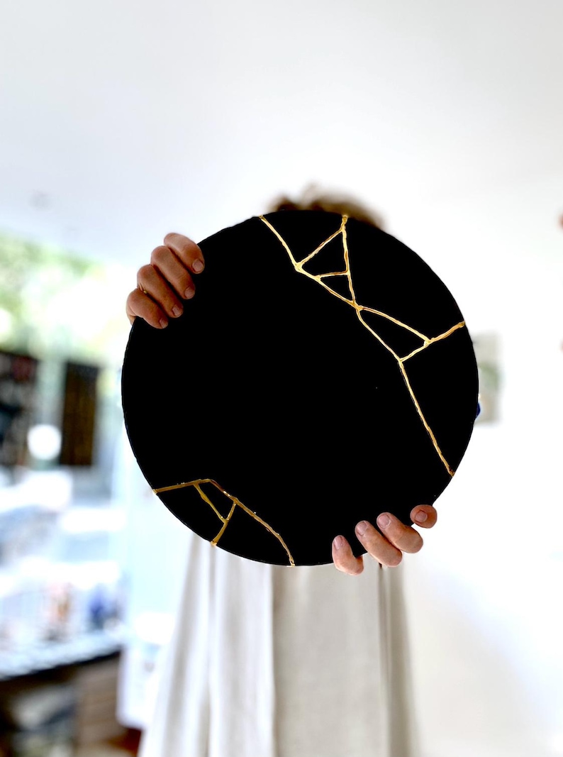Puede incluir: Una pieza decorativa redonda negra con l&iacute;neas doradas al estilo kintsugi. La pieza est&aacute; sostenida por dos manos, y el fondo est&aacute; borroso.