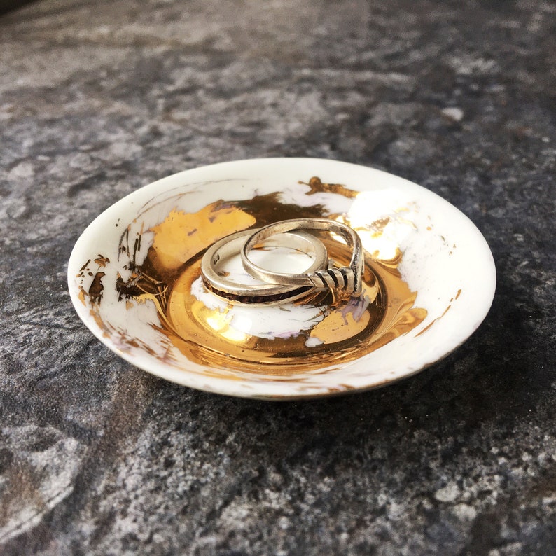 K&ouml;nnte beinhalten: Eine wei&szlig;e Keramik-Ring Schale mit goldenen Akzenten. Die Schale hat einen Ring mit einem gedrehten Design auf einem einfachen Band.