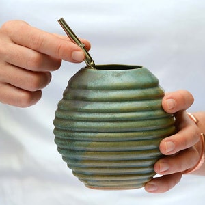 May include: A green ceramic gourd with a ribbed texture, used for drinking mate, a traditional South American beverage. A metal bombilla straw is inserted into the gourd.