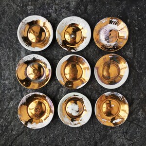 K&ouml;nnte beinhalten: Neun gold- und wei&szlig;farbene Keramikteller mit einem metallischen Schimmer. Die Teller sind rund und haben einen leicht erh&ouml;hten Rand. Die Teller sind in einem Gittermuster auf einer dunkelgrauen Oberfl&auml;che angeordnet.