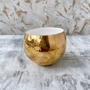 May include: A gold-plated ceramic bowl with a white interior. The bowl is round and has a reflective surface.