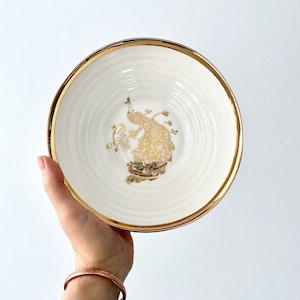 Può includere: Una ciotola in ceramica bianca con un bordo dorato e un design di pavone dorato. La ciotola è perfetta per servire spuntini o dessert.
