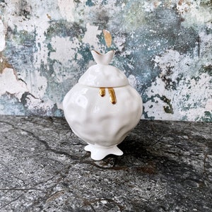 May include: White ceramic jar with a textured surface and gold accents. The lid has a bird-shaped finial. The jar is sitting on a grey surface.