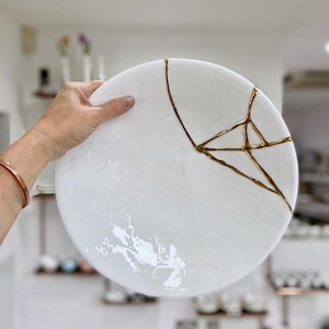 Puede incluir: Un plato de cer&aacute;mica blanco con grietas reparadas con laca dorada. El plato redondo es sostenido por una mano, mostrando el arte japon&eacute;s Kintsugi. La superficie del plato tiene un sutil efecto veteado. Di&aacute;metro aproximado de 25 cm.