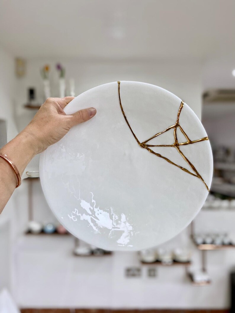 Puede incluir: Un plato de cer&aacute;mica blanco con grietas reparadas con laca dorada. El plato redondo es sostenido por una mano, mostrando el arte japon&eacute;s Kintsugi. La superficie del plato tiene un sutil efecto veteado. Di&aacute;metro aproximado de 25 cm.