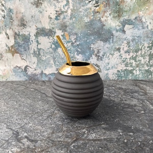 May include: Black ceramic mate gourd with a gold rim and a gold metal bombilla straw. The gourd has a ribbed texture.