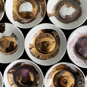 K&ouml;nnte beinhalten: Ein Set aus neun wei&szlig;en Keramik-Schalen mit goldenen und violetten Akzenten. Die Schalen haben ein einzigartiges, handgemaltes Design mit einer metallischen Oberfl&auml;che.