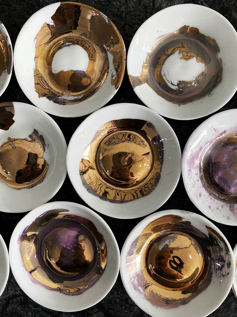 K&ouml;nnte beinhalten: Ein Set aus neun wei&szlig;en Keramik-Schalen mit goldenen und violetten Akzenten. Die Schalen haben ein einzigartiges, handgemaltes Design mit einer metallischen Oberfl&auml;che.