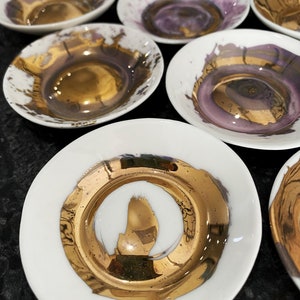 K&ouml;nnte beinhalten: Set aus kleinen wei&szlig;en Keramikschalen mit goldenen Akzenten. Die Schalen haben ein einzigartiges, abstraktes Design mit einem goldenen Rand und einer goldenen Mitte.