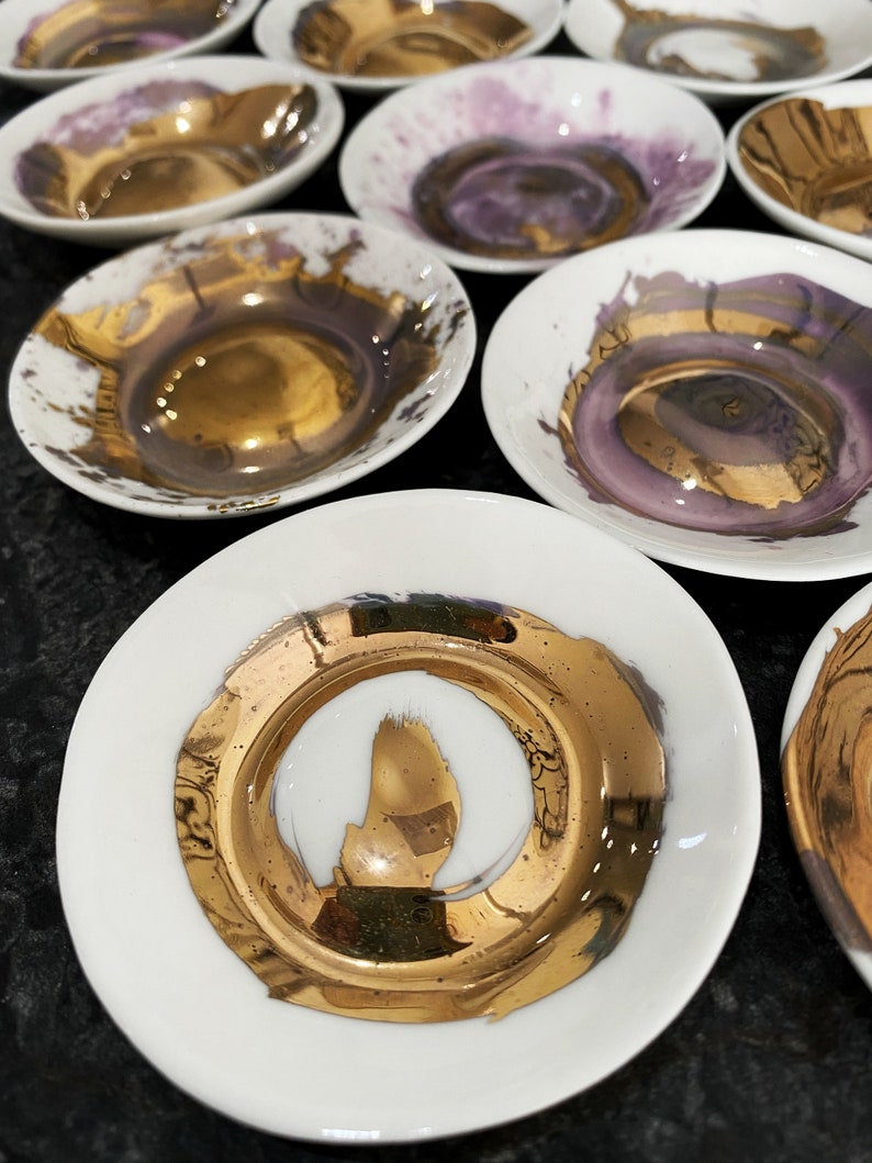 K&ouml;nnte beinhalten: Set aus kleinen wei&szlig;en Keramikschalen mit goldenen Akzenten. Die Schalen haben ein einzigartiges, abstraktes Design mit einem goldenen Rand und einer goldenen Mitte.