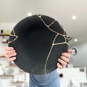 Puede incluir: Un plato redondo de cer&aacute;mica negra con l&iacute;neas doradas de reparaci&oacute;n estilo kintsugi. El plato se sostiene frente a una persona. La superficie del plato es mate y las l&iacute;neas doradas crean un contraste llamativo.