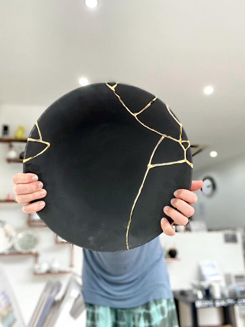 Puede incluir: Un plato redondo de cer&aacute;mica negra con l&iacute;neas doradas de reparaci&oacute;n estilo kintsugi. El plato se sostiene frente a una persona. La superficie del plato es mate y las l&iacute;neas doradas crean un contraste llamativo.