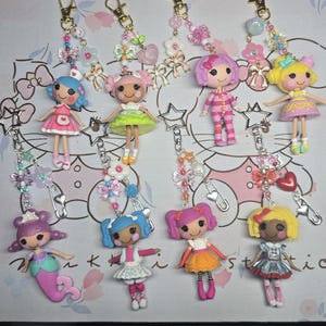 May include: A collection of eight doll keychains. Each doll has a unique outfit and hairstyle, with colors including pink, blue, yellow, and purple. The keychains feature decorative beads, charms, and star-shaped accents, attached to gold-colored clasps.