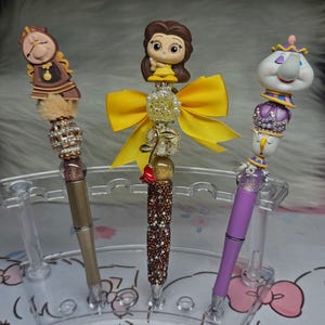 May include: Three decorative pens featuring Beauty and the Beast characters. One pen has a brown and gold clock topper, another has a Belle figurine with a yellow bow, and the third has a Mrs. Potts topper. The pens are displayed in a clear plastic holder.