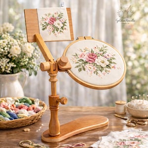 May include: A wooden embroidery stand with a square and round hoop, both featuring floral embroidery with pink and white roses. The stand is made of light wood and has adjustable joints. The Golden Needle Craft Co. logo is visible.