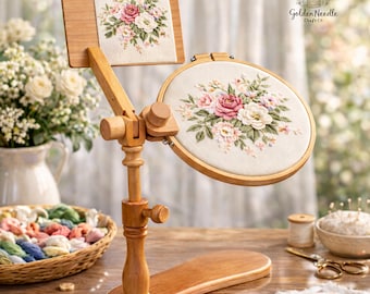 May include: A wooden embroidery stand holds a square and round embroidery hoop, each displaying a floral design with pink and white roses. The stand is made of light-colored wood and has adjustable joints. The Golden Needle Craft Co. logo is visible.