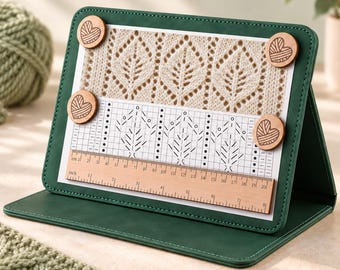 May include: A dark green foldable stand holding a white paper with a beige knit pattern and a black and white knitting chart. Four round wooden magnets with heart-shaped yarn ball engravings secure the paper. A wooden ruler shows 8 inches.