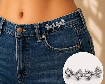 May include: A silver-toned metal waist tightener featuring two connected bow-shaped clips. The accessory is shown attached to the waistband of dark blue denim jeans, with a circular inset providing a clear view of the metallic bow design.