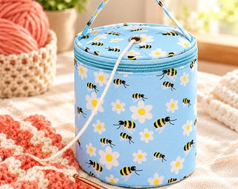 May include: A cylindrical light blue yarn storage bag featuring a repeating pattern of yellow and black bees and white daisies. The bag has a zippered lid with a metal grommet for yarn feeding, a fabric handle, and a single strand of white yarn.