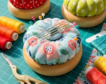 May include: Three round fabric pincushions with wooden bases, featuring red polka dot, green floral, and light blue floral patterns. The pincushions are adorned with colorful sewing pins and needles, accompanied by spools of thread and gold-toned scissors.