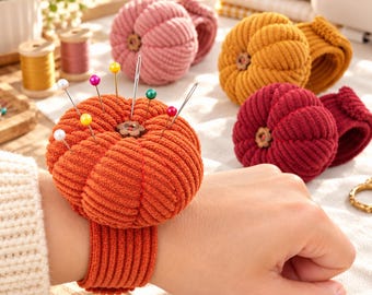 May include: A pumpkin-shaped orange corduroy pincushion with a ribbed texture and a small wooden button center, attached to a matching elastic wrist strap. Several sewing pins with colorful round heads are inserted into the fabric surface.