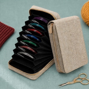 May include: A beige fabric accordion-style storage case with a gold zipper, containing multiple circular knitting needle sets in various colors including purple, teal, red, blue, green, and silver, organized within black interior slots.