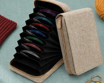 May include: A beige fabric accordion-style storage case with a gold zipper, containing multiple circular knitting needle sets in various colors including purple, teal, red, blue, green, and silver, organized within black interior slots.