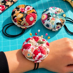 May include: Three round, fabric-covered pincushions featuring floral patterns in black, light blue, and white. Each cushion has a central white bead, white lace trim, and a black elastic wrist strap. One cushion is shown worn on a wrist with pins.